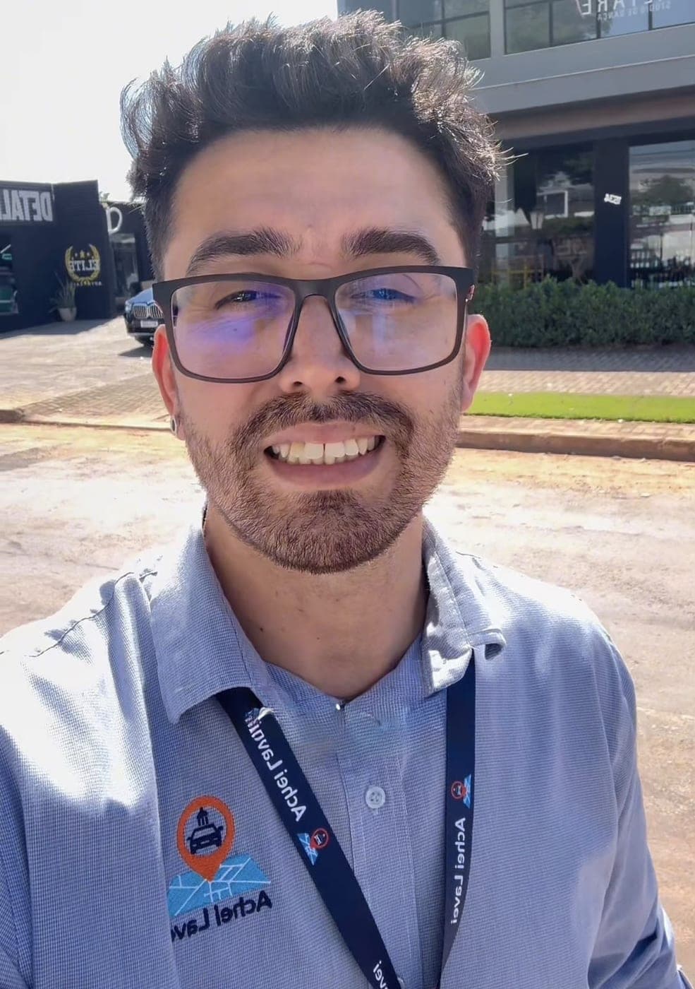 Integrante da equipe Achei Lavei, de camisa com o logotipo bordado e crachá, em frente a uma loja de estética automotiva